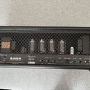 Zadní strana kytarového zesilovače v černém provedení s kovovou mřížkou, pod kterou jsou vidět čtyři elektronky a transformátory. Na černém spodním p…