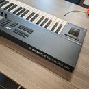 Černý MIDI kontrolér s klávesnicí, umístěný na dřevěném stole. Na boku je bílý nápis KOMPLETE KONTROL S61 s logem výrobce, stejné logo a nápis KOMPLE…