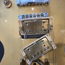 Detail elektrické kytary v zlaté barvě s kovovým hardwarem. Jsou vidět dva snímače typu humbucker v krémových rámečcích, kobylka a strunový most. Pov…