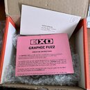 V kartonové krabici s červenými okraji leží růžový manuál s nápisy EXO, GRAPHIC FUZZ a -OPERATING INSTRUCTIONS-. Pod manuálem je průhledná bublinková…