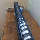 Černá lineární LED lampa s řadou kulatých čoček v plastovém nebo kovovém těle. Zařizení leží na dřevěném stole a je připojeno k napájecímu kabelu. Na…