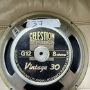 Fotografie zachycuje zadní stranu reproduktoru s kovovým rámem. Na středovém černém štítku je zlatým písmem nápis CELESTION LOUDSPEAKERS, dále označe…