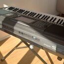 Šedý digitální klavír na X-stánu. Na levé straně je logo YAMAHA a vpravo nápis PSR-S970. Nástroj má ovládací panely s tlačítky,točítky a displejem. P…