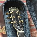 Černá hlava kytary s bílým vlnitým lemováním a zlatými ladičkami. V horní části je bílý nápis GRETSCH a v dolní části bílý podpis Brian Setzer. Nástr…