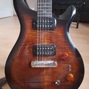 Elektrická kytara s tmavě hnědým průsvitným lakem a viditnou strukturou dřeva. Nástroj má dva černá humbuckery, stříbrný most a dva oranžové oválné k…
