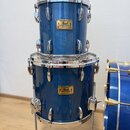 Dvě modré bubny v modrém lakovaném provedení, umístěné jeden nad druhým. Oba mají zlaté štítky s nápisy „Pearl“ a „Masters Studio“. Na horních obručí…