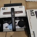 Černý kondenzátorový mikrofon s mřížkou v horní části a přepínačem na těle. Vedle mikrofonu je černá větrná clona a černý pouzdro s vyraženým nápisem…