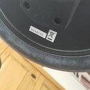 Detail černé plastové části hudebního zařízení. Na povrchu je bílá nálepka s černým nápisem NX46233 a vedle ní symbol přeškrtnuté popelnice. Povrch p…