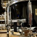 Detailní pohled na černý snare drum s chromovaným kováním. Na horním obruči je viditelný nápis "Mich". Nástroj má připevněný černý textilní popruh s…