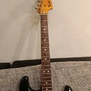 Černá elektrická kytara s hnědým hmatníkem a bílým pickguardem. Na hlavě nástroje je vidět logo Fender® STRATOCASTER. Povrch kytary vykazuje známky o…