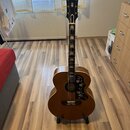 Akustická kytara v hnědé barvě s tmavým pickguardem zdobeným květinovým motivem. Na hlavě hmatníku je bílý nápis Epiphone. Kytara stojí na černém sto…