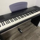 Černý digitální piano s bílými a černými klávesami, umístěné na přívstupném černém stojanu. Na levé straně horního panelu je bílý nápis KURZWEIL. Nás…