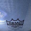 Detail průhledné bubny na buben. Na povrchu je černý nápis REMO v korunovitém rámu a pod ním text AMBASSADOR CLEAR. Povrch vykazuje drobné škrábance…