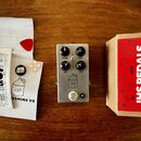 Stříbrný efekt pedál s nápisem JHS PEDALS a XXX doprovázený ikonou kanystru. Na pedálu jsou čtyři černé oválné knoflíky s popisky VOLUME, DRIVE, TONE…