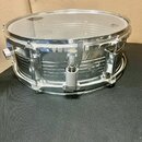 Stříbrný kovový snare drum s průhlednou blánou. Nástroj nemá žádné viditelné značky ani nápisy. Povrch kovu vykazuje drobné škrábance a známky opotře…