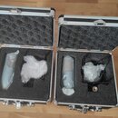 Dvě otevřené hliníkové transportní kufry s černou pěnovou výplní. V každém kufru se nachází jeden světle modrý mikrofon zabalený v průhledné bublinko…