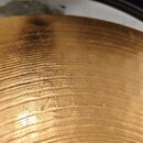 Detail zlatavého kovového povrchu s koncentrickými rýhami. V centru je vyryt nápis „PAISTE“ a pod ním označení „24"“. Povrch vykazuje drobné škrábanc…