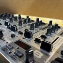 Detailní pohled na horní panel stříbrného DJ mixeru s mnoha černými otočnými knoflíky a posuvníky. Na povrchu jsou viditelné nápisy „CURVE“, „EQ BYPA…