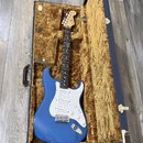 Modrá elektrická kytara typu Stratocaster s bílým pickguardem, uložená v otevřeném tmavomodrém kufru s hnědým plyšovým vnitřkem. Na hlavě kytary je v…