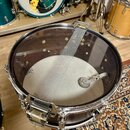 Fotografie znázorňuje snare drum s průhledným plastovým krytem. Na krytu je viditelný nápis REMO CLEAR CORNERS MADE IN USA. Nástroj má chromované obr…