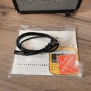 V průhledném plastovém sáčku se nachází černý napájecí kabel s vidlicí a dokumentace. Na přiloženém červeném kousku papíru je bílé logo Fender a nápi…