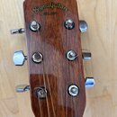 Fotografie zachycuje hlavu akustické kytary z hnědého dřeva s chromovanými ladičkami. V horní části je žlutý nápis Sigma Guitars a pod ním text EST.…