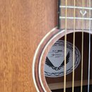 Detail akustické kytary s dřevěným povrchem a viditelným hmatníkem. Uvnitř ozvučnice je bílá štítková nálepka s nápisy CAL INSTRUMENTS, FLY MAH MODEL…