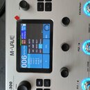 Stříbrný efektový procesor s nápisem M-WAVE a označením 300 Effects Processor. Uprostřed je zapnutý displej s modrým rozhraním, kde jsou vidět nápisy…