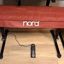 Červený textilní kryt s bílým nápisem nora zakrývá klavír nebo syntezátor na černém kovovém stojanu. Na podlaze je umístěn černý sustain pedál s malý…