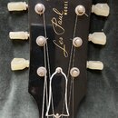 Černá hlava kytary s bílým logem Gibson a zlatým nápisem Les Paul MODEL. Na hlavě jsou vidět šest ladicích kolíčků s krémovými knoflíky. Povrch vykaz…