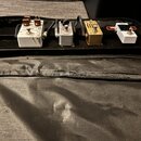 Čtyři kytarové efekty umístěné na černém pedalboardu v černé přepravní tašce. Zleva jsou vidět bílý pedál s hnědými knoflíky, stříbrný pedál s nápise…