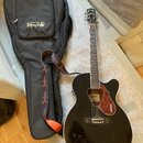 Černá akustická kytara s vyřezem v těle a hnědým rýhovaným plektrem. Na hlavě nástroje je nápis GRETSCH a na plektru jsou dvě loga GRETSCH. Kytara je…