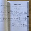 Fotografie zachycuje otevřenou knihu s notovým zápisem a tabulaturami pro kytaru. Stránka nese nadpis PRIMITIVE BLUES-03 a obsahuje text v češtině, a…