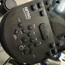 Černý elektronický modul k bubnům s nápisem NITRO DRUM MODULE. Na panelu jsou umístěna gumová ovládací tlačítka s popisky jako RECORD, CLICK, DRUM OF…