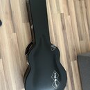 Černý tvrdý kufýr na kytaru s rukojetí. Na povrchu jsou dvě bílá loga ve tvaru kosočtverců s nápisy „Epiphone“ a „Gibson“. V horní části těla je umís…