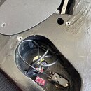 Fotografie zachicuje zadní stranu černé kytary s otevřeným přístupovým otvorem do elektroniky. Uvnitř jsou vidět černé kabely, součástky a červený ko…
