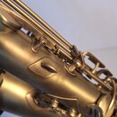 Detailní záběr na část saxofonu v matném zlatavém povrchu. Jsou vidět mechaniky, hroty a spojovací prvky nástroje. Na povrchu nejsou žádné čitelné zn…