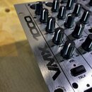Detailní pohled na stříbrný mixovací pult s kovovým povrchem. Na boku je černý nápis VMX 1000. Jsou vidět řady černých otočných ovladačů s bílým znač…