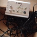 Stříbrný ovládací modul s nápisem Roland a DRUMS. Na přední straně jsou čtyři bílé otočné ovladače s popisky VOLUME a TEMPO, pět číslovaných tlačítek…