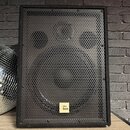 Černý reproduktor v bedně potažené tmavým materiálem s kovovou mřížkou. Na přední straně je umístěn malý bílý čtverečkový štítek s nápisem the box. P…