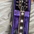 Černá elektrická kytara v otevřeném kufru s fialovou výstuhou. Na hlavě hmatníku je bílé logo Epiphone a pod ním symbol v podobě pěstního uzlu. Hmatn…