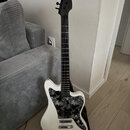 Bílá elektrická kytara s černým hmatníkem a černou hlavou. Kytara má černý pickguard s bílým květinovým vzorem a dva snímače typu single-coil. Na nás…