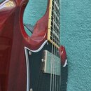 Detailní pohled na červenou elektrickou kytaru s tmavým hmatníkem a krémovým bindingem. Nástroj má černý pickguard s bílým lemováním a jeden viditeln…