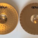 Dvě mosazné činely s logem Paiste. Levý činel má povrch zdobený řadami důlků, pravý činel má po obvodu vyřezané zářezy. Oba nástroje vykazují mírné z…
