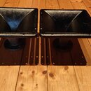 Dvě černé tubové reproduktory s hranatým hrdlem, umístěné na dřevěné podlaze. Povrch je černý s mírným leskem a vykazuje drobné známky opotřebení. Na…