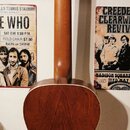 Zadní pohled na akustickou kytaru s hnědým dřevěným povrchem a světlým lemováním kolem korpusu. Nástroj je umístěn v nástěnném držáku. Na hlavě kytar…