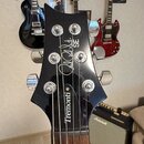 Černá hlava elektrické kytary s nápisy Paul Reed Smith SE a Tremonti. Jsou vidět chromované ladičky a struny. V pozadí jsou další kytary, Marshall ze…