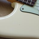 Detailní záběr na tělo a hриф elektrické basové kytary v krémové barvě. Nástroj má světle zelený pickguard a hриф z tmavého dřeva s bílými tečkovaným…