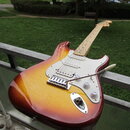 Elektrická kytara s tělem v barvě sunburst a bílým pickguardem. Na hlavě hředu je viditelné logo Fender. Nástroj má tři snímače, tři ovládací knoflík…