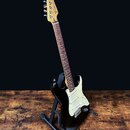 Černá elektrická kytara typu Stratocaster s bílým pickguardem a hnedým hmatníkem. Na hlavě nástroje je viditelné logo Fender. Kytara je umístěna na č…